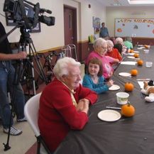 Photo of elderly woman working with young student on art project with pumpkins and a video camera in the background