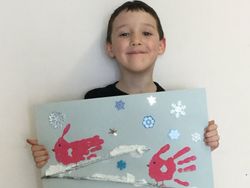 Picture of a student holding up his artwork of a winter scene made with red handprints to look like birds with snowflakes and a blue background