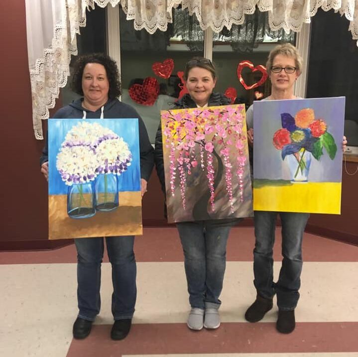 Photo of three ladies holding up flower theme wine and canvas paintings