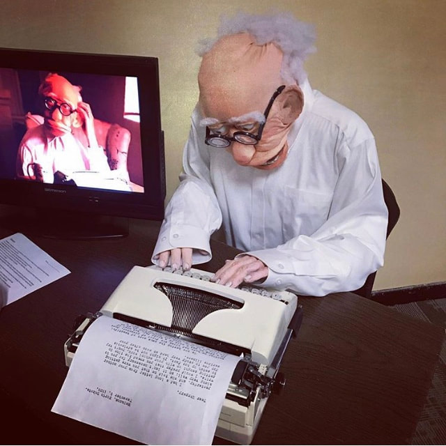 Photo of art exhibit display of a puppet dressed as an old man typing on a typewriter